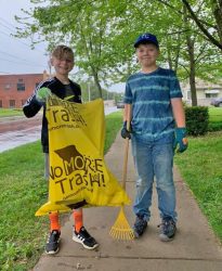 two boys pose with a branded trash bag and rake while cleaning up a neighborhood